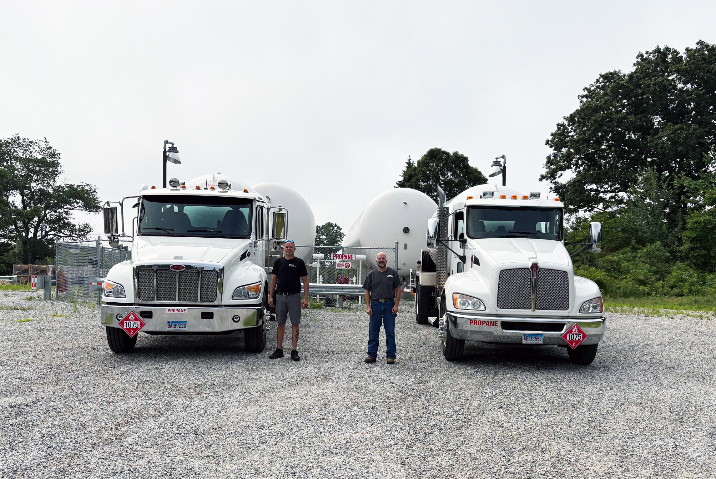 Two men stand between two white semi-trucks parked on a gravel lot at the Daniels propane facility, with large tanks and trees visible in the background.