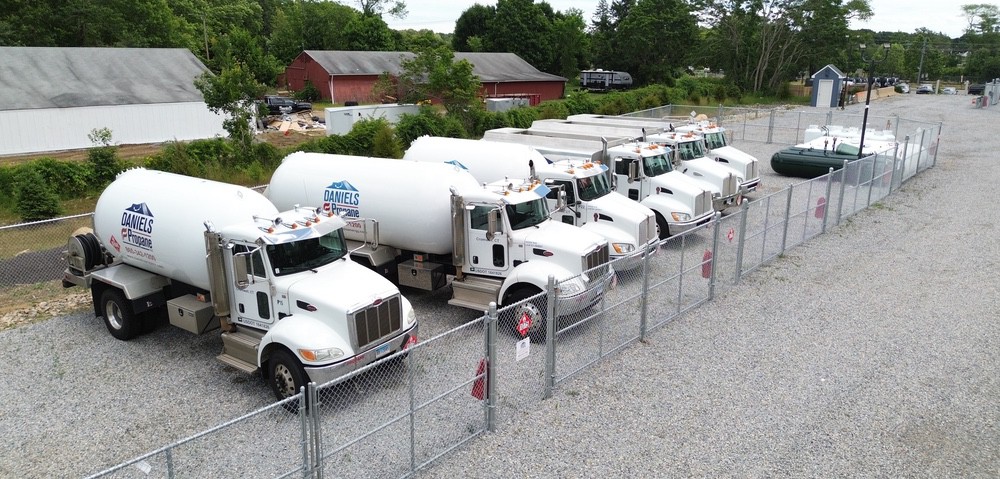 A row of six white Daniels Energy propane delivery trucks is parked inside a fenced lot on a gravel surface, ready to meet your propane and heating oil needs.