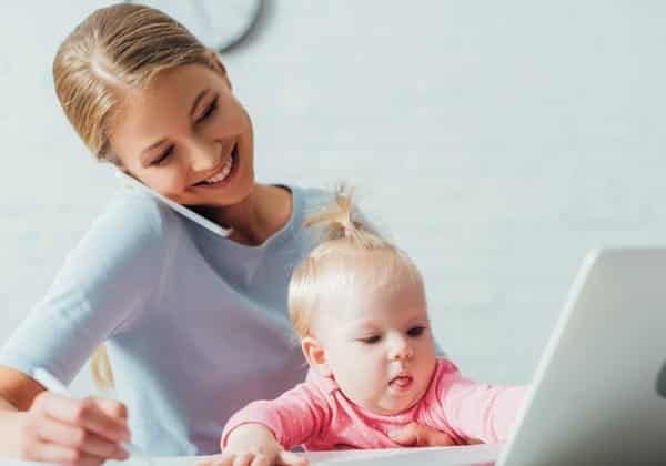 A woman multitasking with a laptop and a baby in front of her.